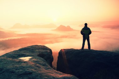 Moment of loneliness. Man on the rock empires  and watch over the misty and foggy morning valley to Sun clipart