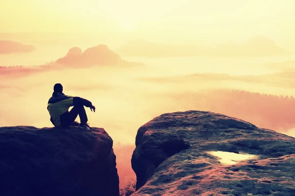 Visão traseira do caminhante masculino sentado no pico rochoso enquanto desfruta de um amanhecer colorido acima do vale das montanhas — Fotografia de Stock