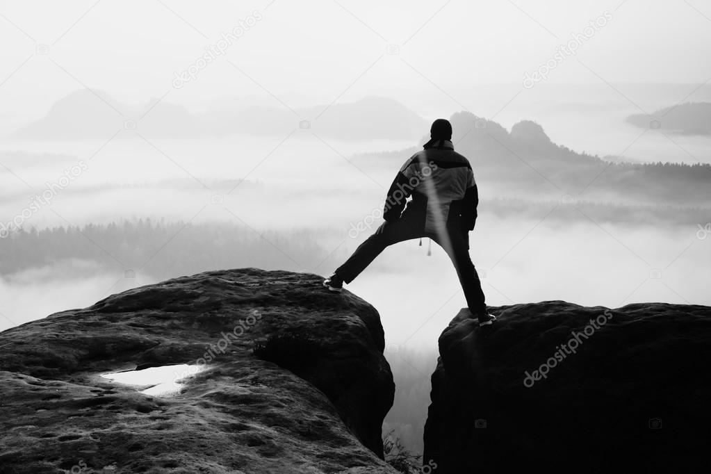 Hiker in black is jumping between the rocky peaks. Wonderful daybreak in rocky mountains, heavy orange mist in deep valley. Miracle of nature