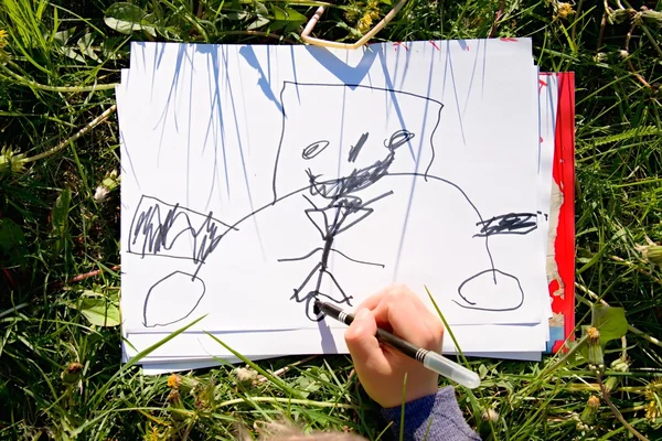 Vista de perto da mão da criança que desenha ornamentos coloridos no papel. Caderno na grama verde fresca do prado . — Fotografia de Stock