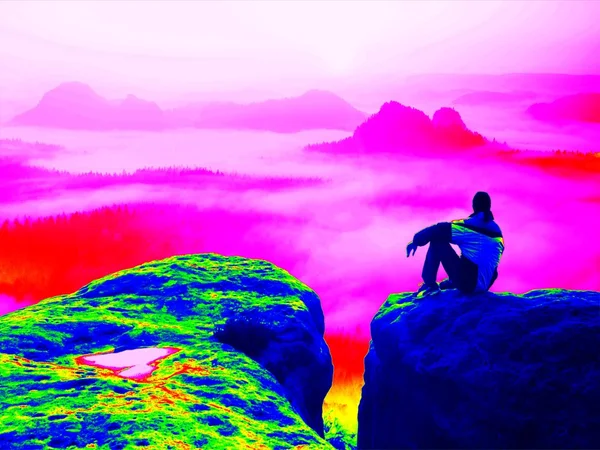 Fantastic infrared scan. Tired tourist on the rocky peak. Wonderful daybreak in mountains, heavy orange mist in deep valle — Stockfoto