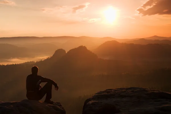 Turista v černý vítr bunda sedět na ostrý kámen a dívat se do barevné mlhavé údolí níže. Slunečné jarní svítání v rocky mountains. — Stock fotografie