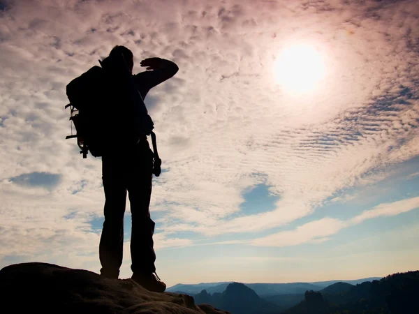 Tourist with big backpack stand on rocky view point and watching into deep misty valley bellow. Sunny spring daybreak in rocky mountains. — Stock Photo, Image