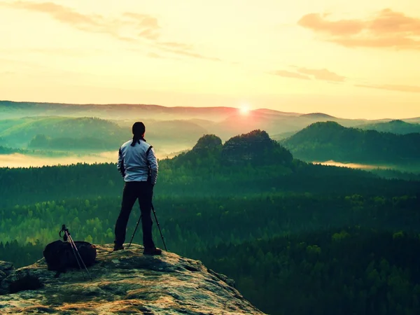 Professional photographer in white jacket  takes photos with camera on tripod on rocky peak. Dreamy fogy landscape, spring orange pink misty sunrise in a beautiful valley below — Stock Photo, Image