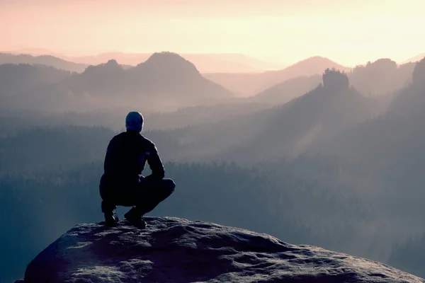 Jeune homme en tenue de sport noire est assis sur le bord de la falaise et regardant vers la vallée brumeuse ci-dessous Photo De Stock