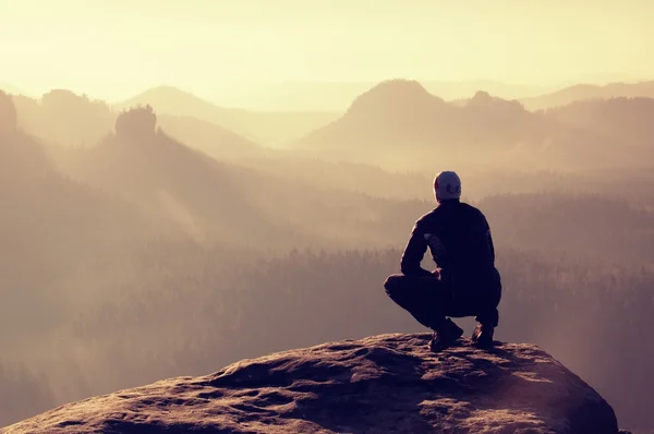 Young man in black sportswear is sitting on cliff edge and looking to misty valley bellow — Stock Photo, Image