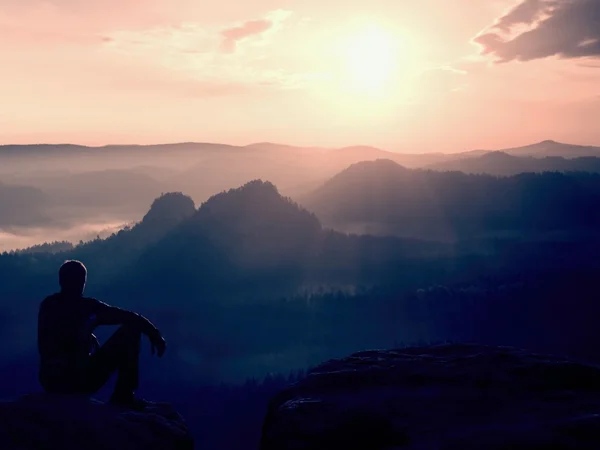 Touristen in schwarzer Windjacke sitzen auf einem scharfen Felsen und blicken in ein buntes Nebeltal. Sonnenaufgang im Frühling in den felsigen Bergen. — Stockfoto