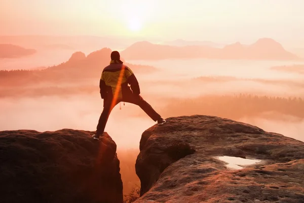 Wandelaar in het zwart is springen tussen de rotsachtige pieken. prachtige daybreak in rocky mountains, zware Oranje mist in diepe vallei. wonder van de natuur — Stockfoto