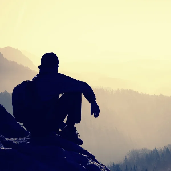 Tired hiker with sporty backpack sit on rocky peak and watching into deep misty valley bellow. Sunny spring daybreak in rocky mountains. — Stock fotografie