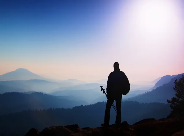 Silhouette of tourist with poles in hand. Hiker with sporty backpack stand on rocky view point above misty valley. Sunny spring daybreak in rocky mountains. — Stock Photo, Image