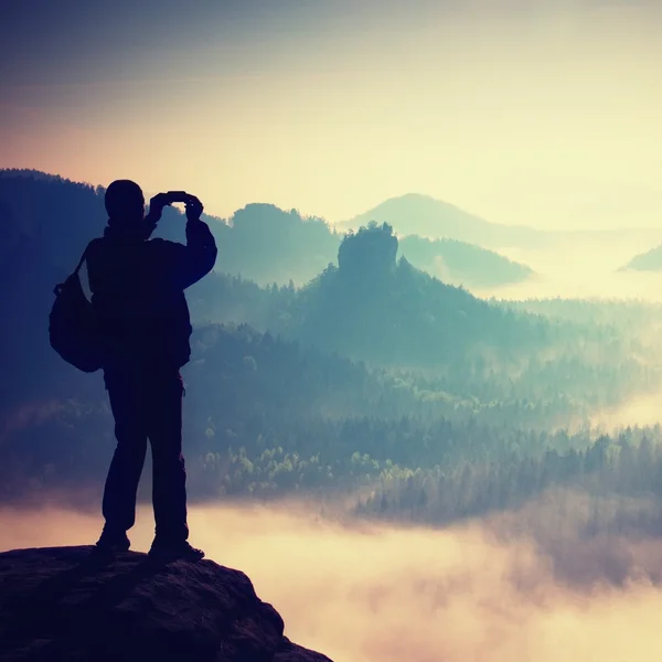 Turista con zaino scatta foto con smartphone sulla vetta della roccia. Valle nebbiosa da sogno sotto — Foto Stock