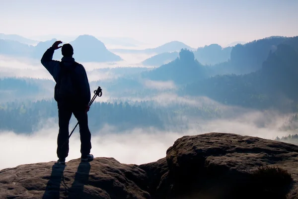 Misty day in rocky mountains. Silhouette of tourist with poles in hand. Hiker stand on rocky view point above misty valley. — Stock Photo, Image