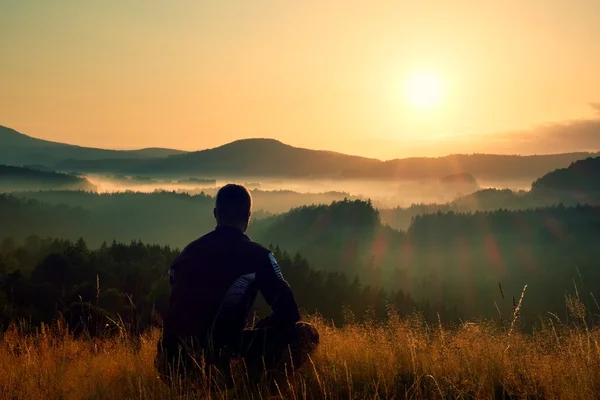 Hiker in squatting position in high grass meadow  enjoy the colorful sunrise scenery — Stok fotoğraf