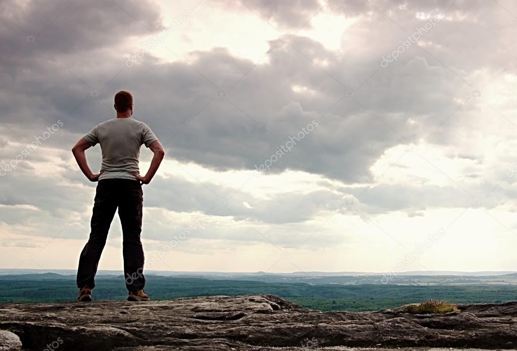 Gesture of triumph. Happy hiker in greyshirt and dark trousars. Tall man on the peak of sandstone cliff watching down to landscape.