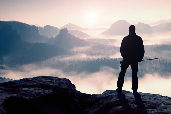 Giorno nebbioso in montagne rocciose. Silhouette di turista con pali in mano. Escursionista in piedi sul punto di vista roccioso sopra la valle nebbiosa . — Foto Stock