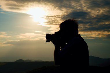 Professional photographer in jeans and shirt  takes photos with mirror camera on peak of rock. Dreamy landscape, orange  Sun at horizon clipart