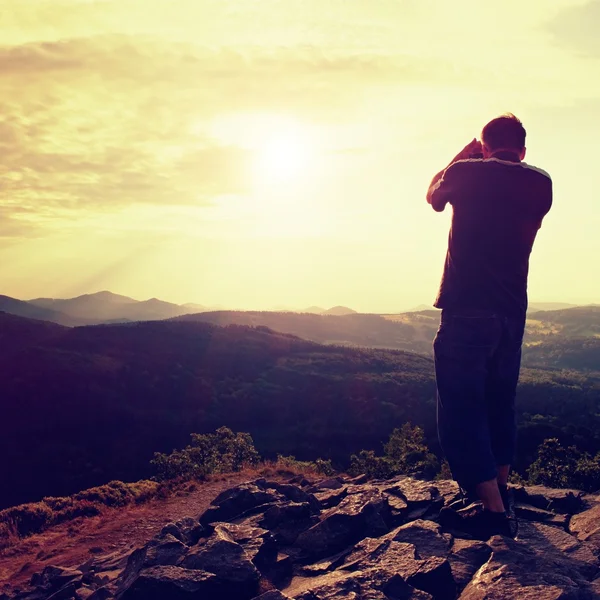 Fotógrafo profesional en jeans y camisa toma fotos con cámara de espejo en el pico de roca. Paisaje de ensueño, Sol naranja en el horizonte —  Fotos de Stock