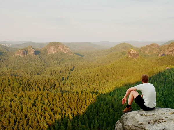 Moment of loneliness. Man in shirt sit on the cliff of rock and watching into forest valley. — Stockfoto