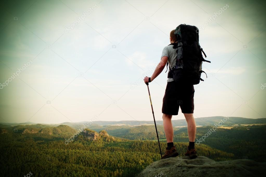 Tall backpacker with poles in hand. Sunny summer evenng in rocky mountains. Hiker with big backpack stand on rocky view point above valley. 