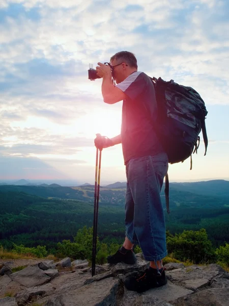 Photographer with big mirror camera on neck and backpack stay on peak of rock. Hilly landscape, fresh green color in valley. — Stock Photo, Image