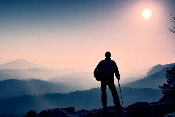 Silhouette of tourist with poles in hand. Hiker with sporty backpack stand on rocky view point above misty valley. Sunny morning in rocky mountains. — Stock Photo, Image