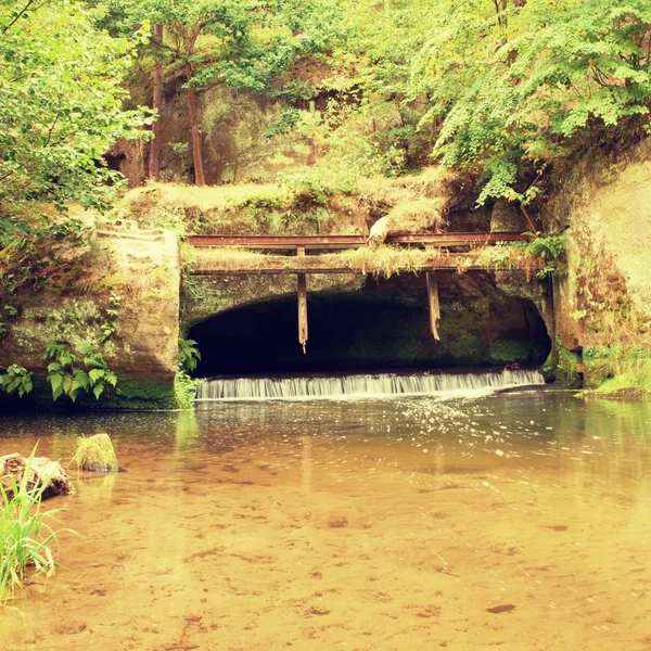 Ein kleines Wehr am Fluss fließt aus der Höhle. Kaltes Wasser eines kleinen Flusses fließt über ein kleines steinernes Wehr. Steiniger und rostiger Bau des Wehrs. — Stockfoto