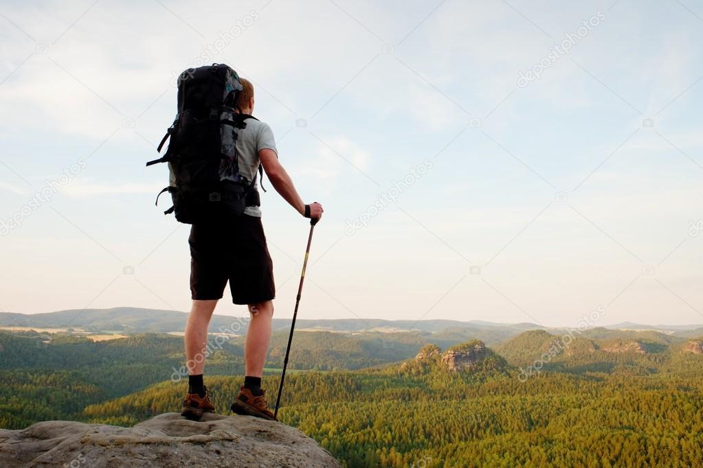 Tall backpacker with poles in hand. Sunny summer evenng in rocky mountains. Hiker with big backpack stand on rocky view point above valley.