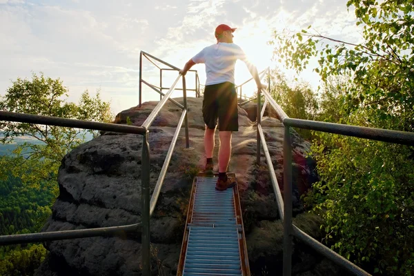 L'escursionista stanco tiene il corrimano in cima. Sunny alba di primavera in montagne rocciose. Escursionista con berretto da baseball rosso, pantaloni scuri e camicia bianca . — Foto Stock