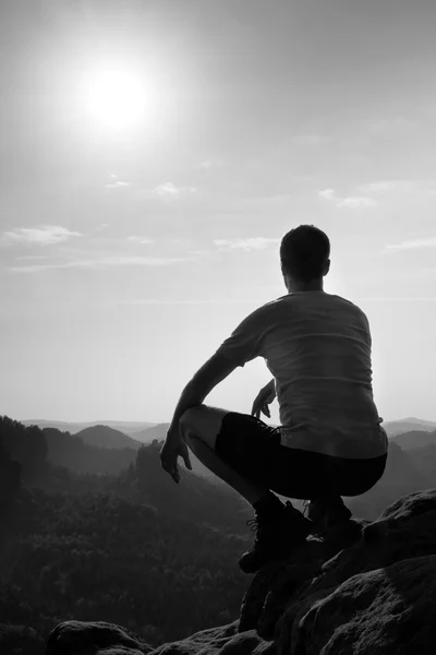 Hombre joven en pantalones deportivos negros y camisa gris está sentado en el borde del acantilado y mirando al valle brumoso abajo —  Fotos de Stock