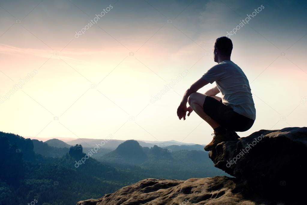 Young man in black sports pants and grey shirt  is sitting on cliff's edge and looking to misty valley bellow