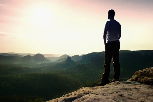 Satisfy tall hiker in grey shirt and dark trousers. Sprtsman on the peak of sharp rock edge  watching down to landscape. — Stock Photo, Image