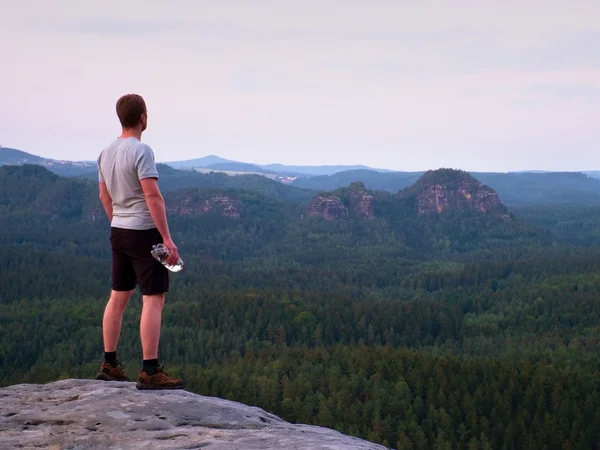 Thirsty hiker in black pants with bottle of water. Sweaty tired tourist on the peak of sandstone rocky park Saxony Switzerland watching into misty landscape.