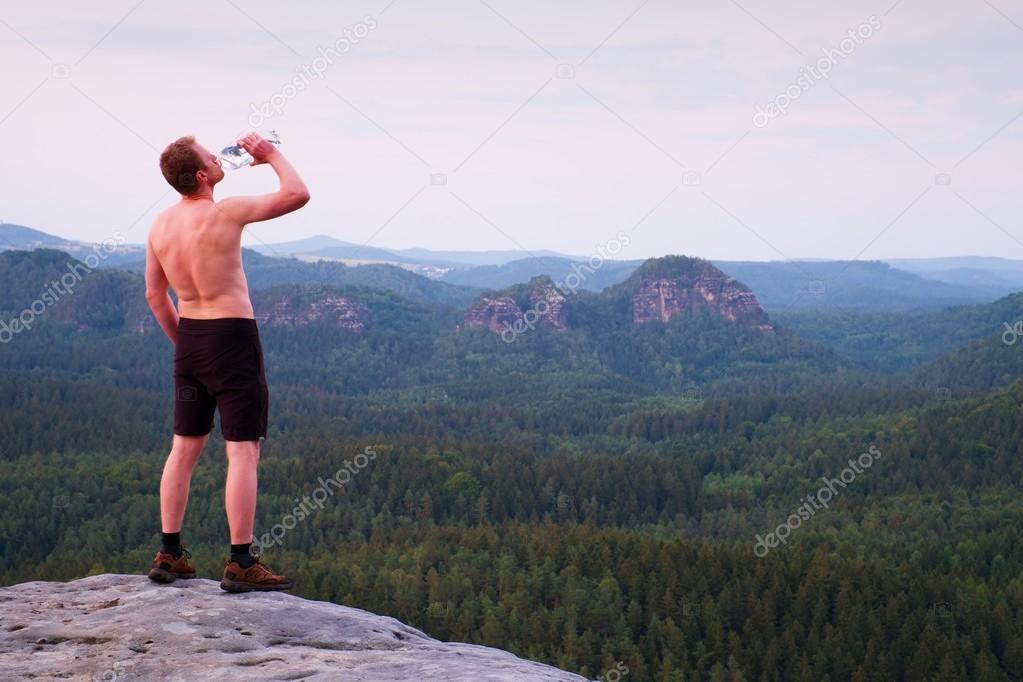 Thirsty hiker in black pants with bottle of water. Sweaty tired tourist on the peak of sandstone rocky park Saxony Switzerland watching into misty landscape.