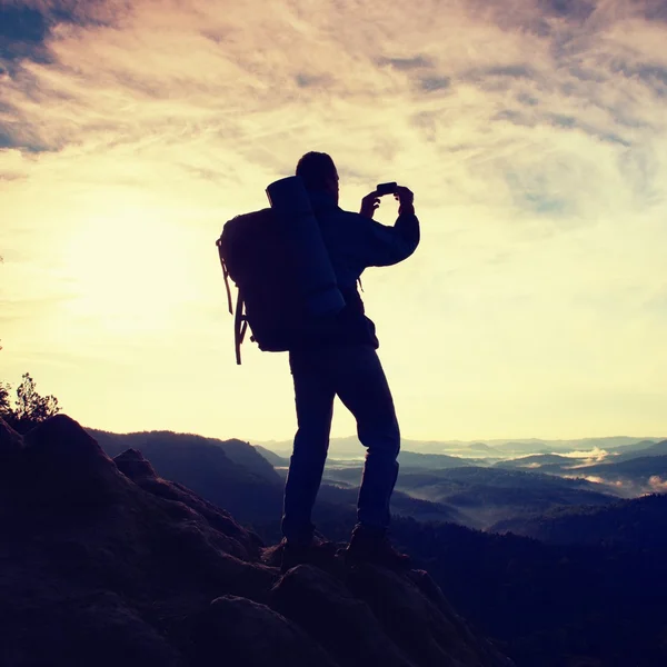 Touristin mit schwerem Rucksack fotografiert mit Smartphone. Mann auf dem felsigen Gipfel. verträumtes Nebeltal unten — Stockfoto