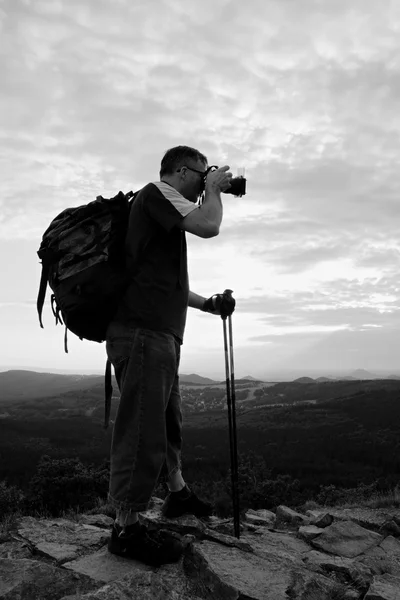 Фотограф с большой зеркальной камерой на шее и рюкзаком остается на вершине скалы. Холмистый пейзаж, свежий зеленый цвет в долине . — стоковое фото