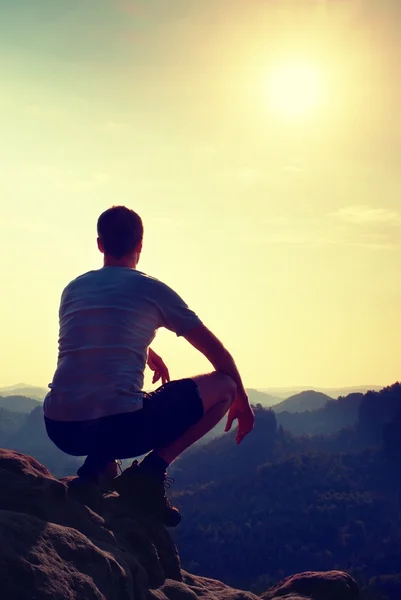 Young man in black sports pants and grey shirt  is sitting on cliff's edge and looking to misty valley bellow — Stock Photo, Image