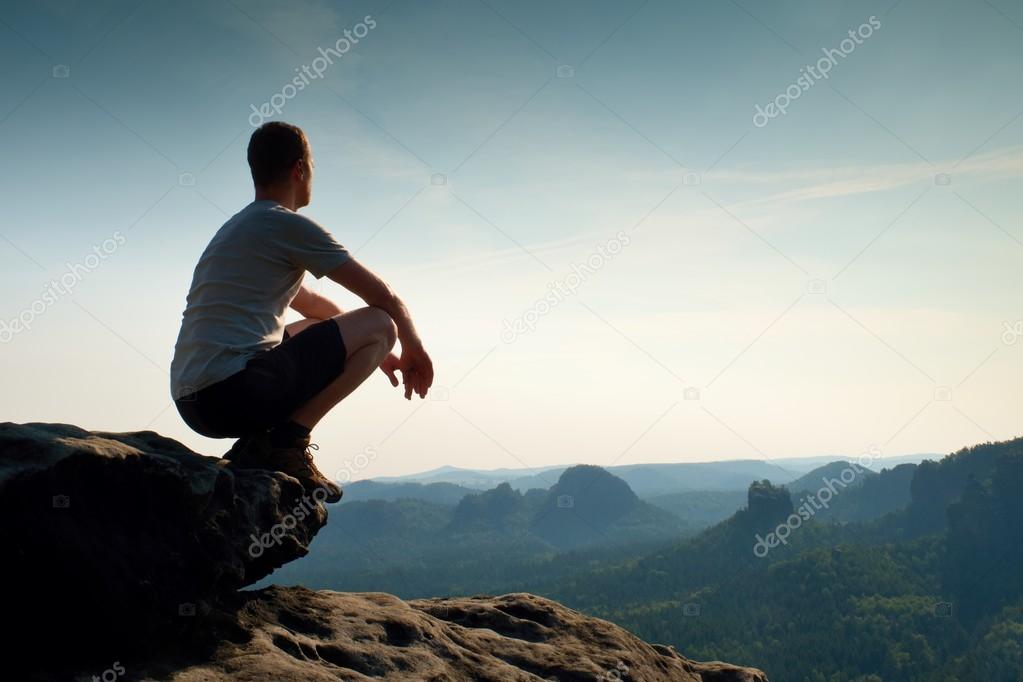 Young man in black sports pants and grey shirt  is sitting on cliff's edge and looking to misty valley bellow