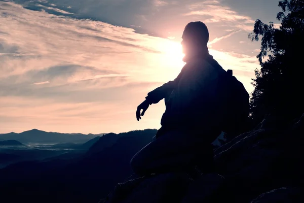 Wandelaar met sportieve rugzak zitten op de rand van de rotsachtige klip en kijken in mistige vallei balg. Zonnige lente daybreak in de rocky mountains. — Stockfoto