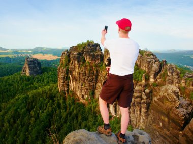 Uçurum rock ve alır fotoğraf peyzaj feryat akıllı telefon kısa saçlı adam. Güneşli bir günde rocky Dağları. Uzun yürüyüşe çıkan kimse gri gömlek ve renkli pantolon.
