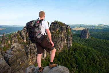 Uçurum rock ve alır fotoğraf peyzaj feryat akıllı telefon kısa saçlı adam. Güneşli bir günde rocky Dağları. Uzun yürüyüşe çıkan kimse gri gömlek ve renkli pantolon.