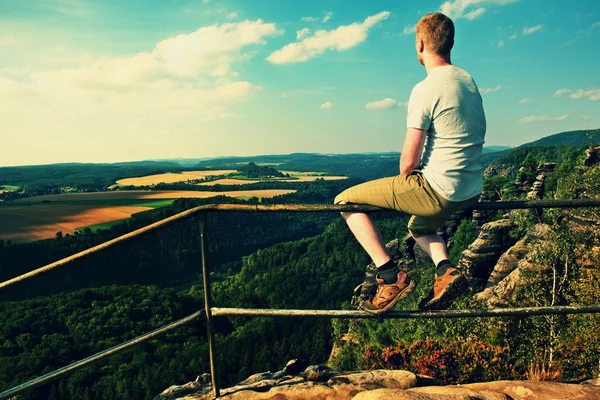 Homme roux assis sur la main courante au sommet de la roche et regarder au paysage. Journée ensoleillée dans les montagnes rocheuses. Randonneur avec chemise, pantalon et bottes gris . — Photo