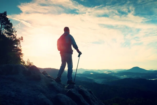 Turist guide med stolpe i handen. Vandrare med sportig ryggsäck stå på rocky synvinkel ovan misty valley. Soliga våren gryning — Stockfoto