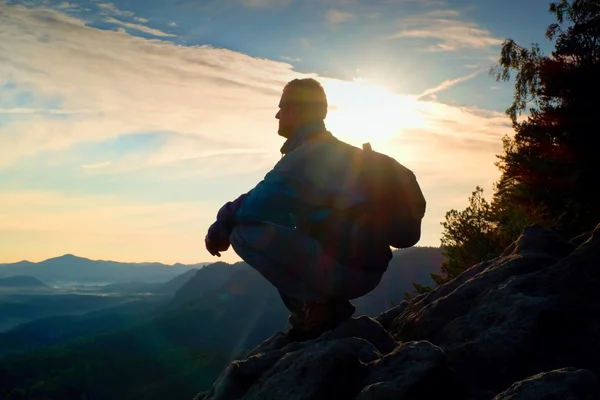 Hiker with sporty backpack sit on rocky cliff edge and watching into misty valley bellow. Sunny spring daybreak in rocky mountains. — Stock Photo, Image