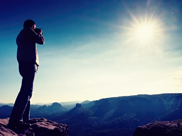 Professional photographer takes photos with mirror camera on cliff of rock. Dreamy misty landscape, hot Sun above — Stock Photo, Image