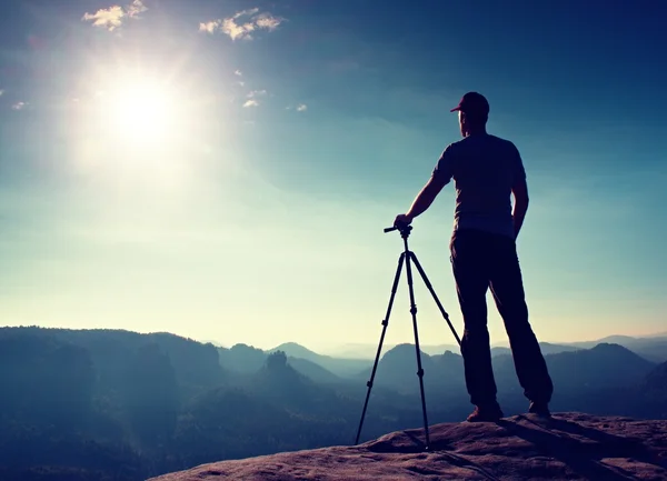 Профессиональный фотограф остается с штативом на скале и мышления. Мечтательный туманный пейзаж, голубой туманный восход солнца в красивой долине внизу — стоковое фото