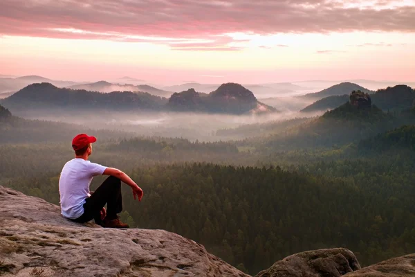 Pria dewasa dengan kemeja putih, trousar gelap dan topi merah. Ginger pria rambut pendek duduk di tebing tajam di atas lembah di taman pegunungan berbatu — Stok Foto