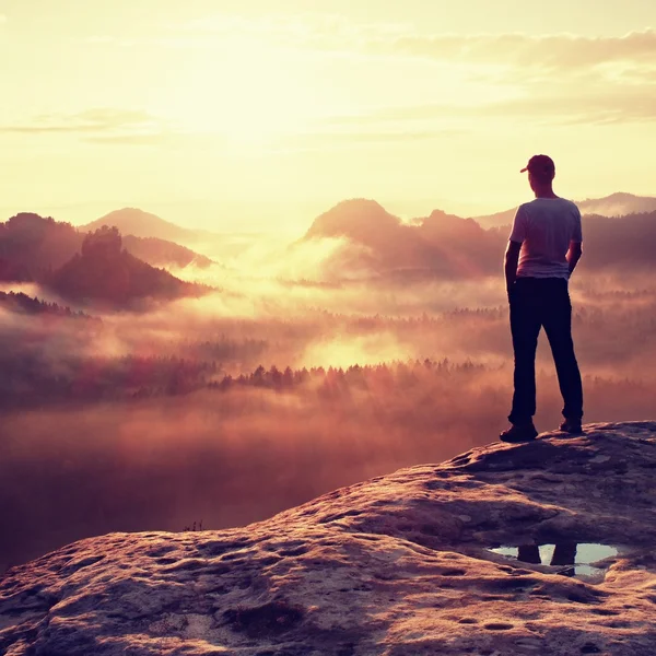 Alone hiker in white shirt and red cap stand on peak of rock in rock empires park and watch over misty and foggy valley to Sun. The miracle of nature — Stock Photo, Image