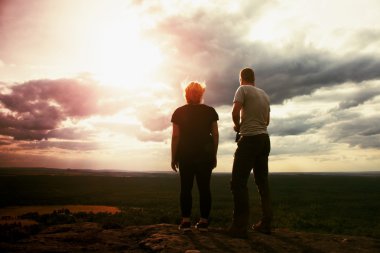 Couple enjoying marvellous moments during sunset . Young pair of hikers on the peak of rock watch over valley to Sun. clipart