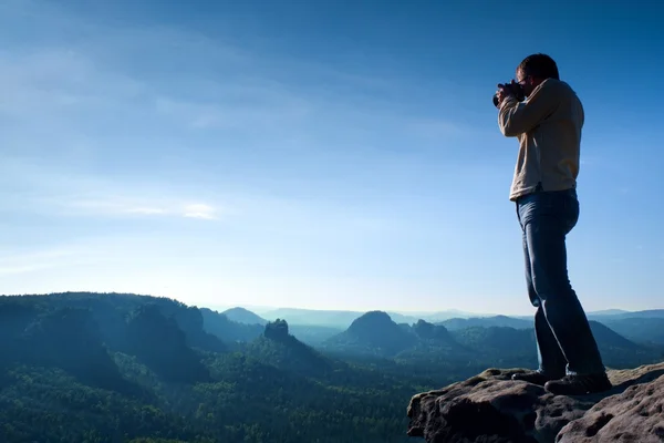 Fotógrafo profissional tira fotos com câmera de espelho no penhasco da rocha. Paisagem nebulosa sonhadora, sol quente acima — Fotografia de Stock