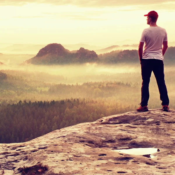 Långe mannen i vit skjorta och svarta byxor med röd keps bo på skarpa klippa och titta till dalen. Färgglada Dimmig morgon i steniga park. — Stockfoto